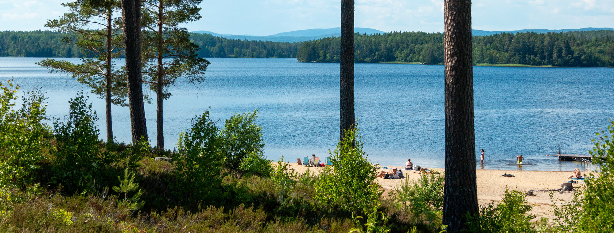 Bilden visar en utsikt mot vatten och en sandstrand med människor som badar.